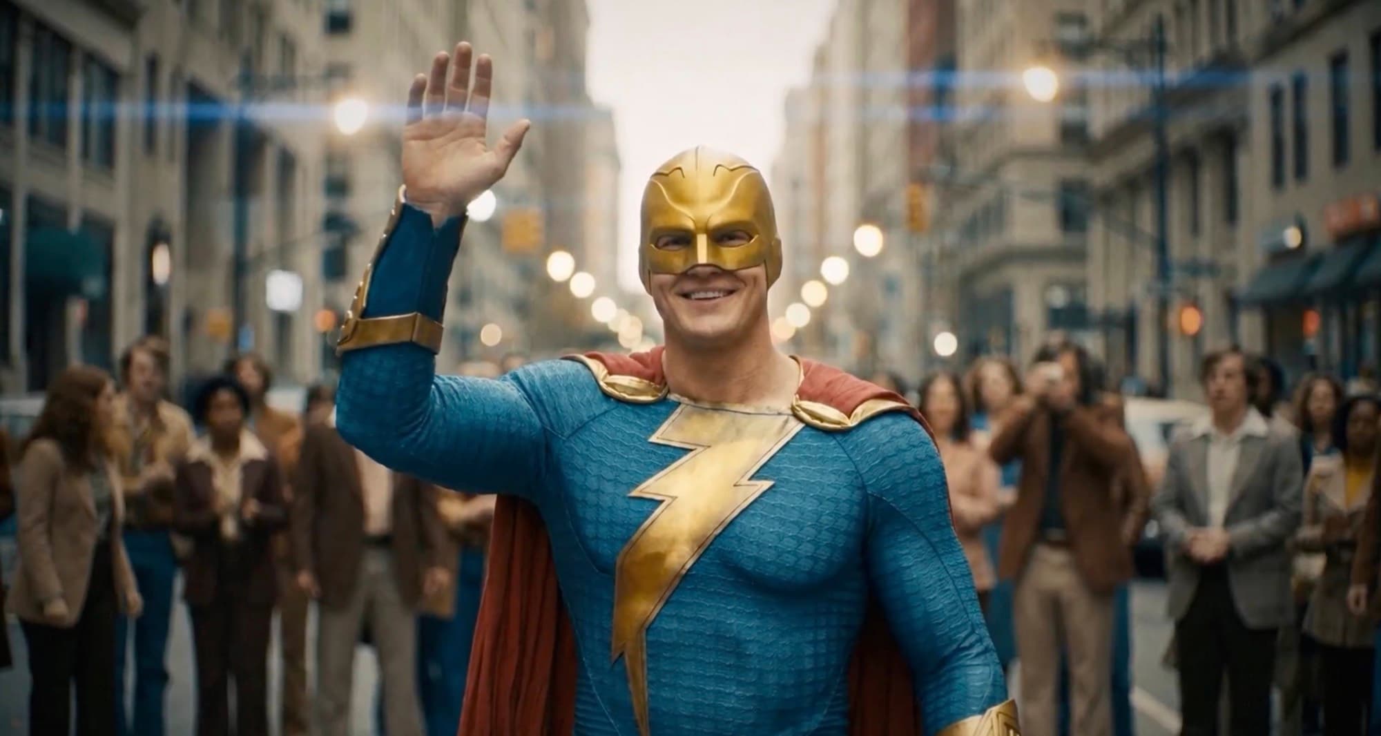 A caped superhero in a gold mask waving on a period city street lined with onlookers.